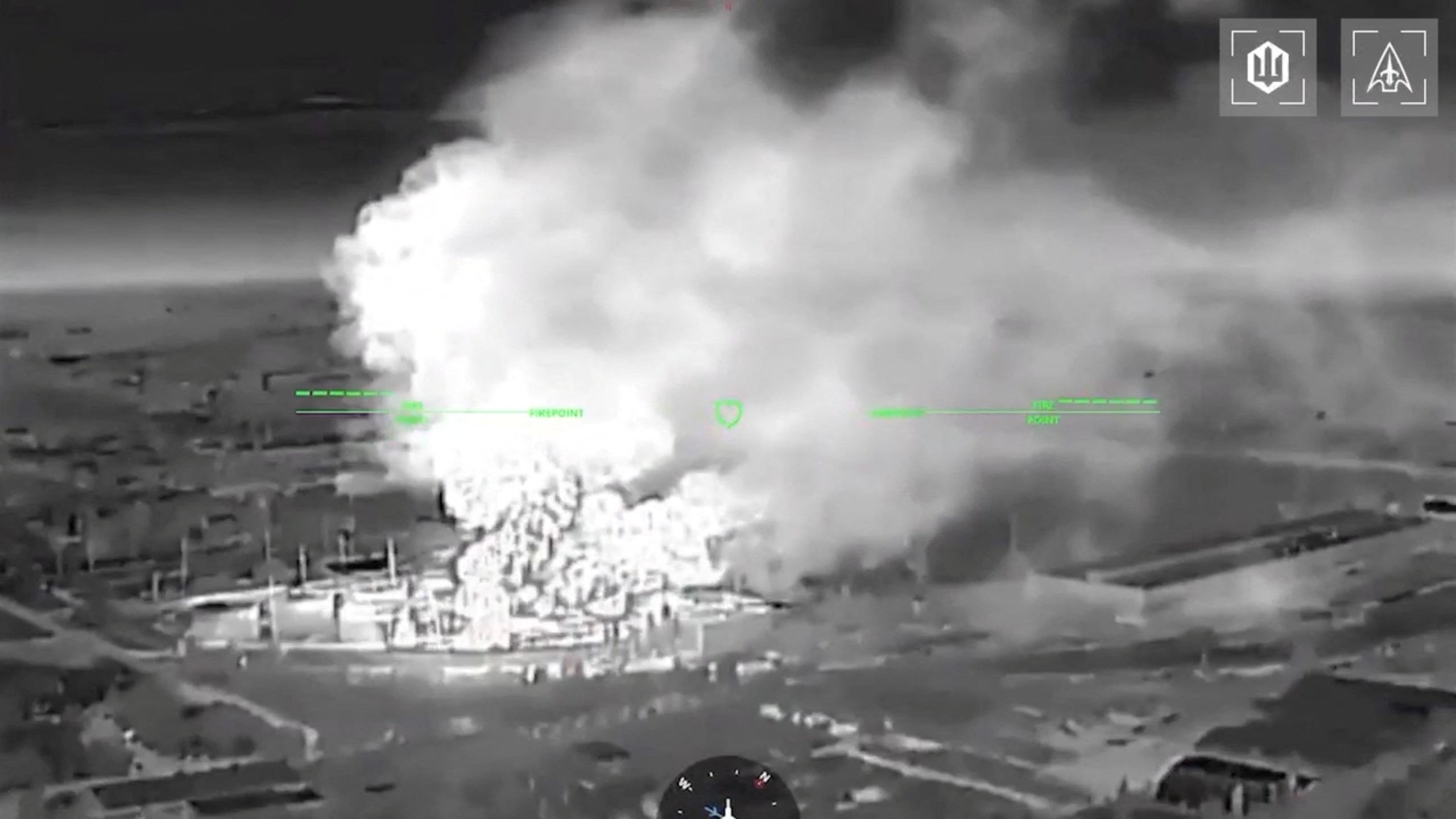 A black and white drone footage shows fire and smoke rising from buildings