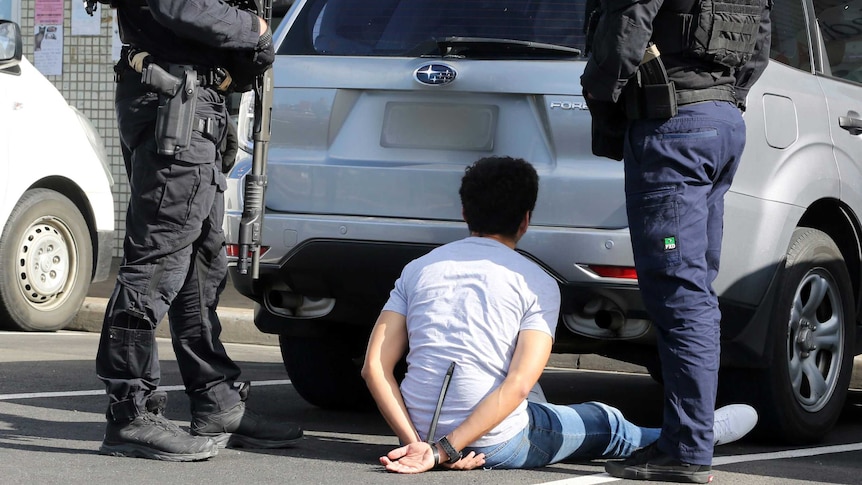 A man sits on the ground behind a car with two armed police standing over him.