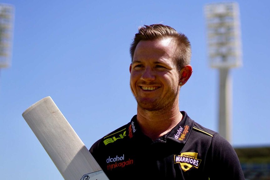 Cricketer D'arcy Short smiles with a bat at the WACA with a blue sky and floodlights in the background.