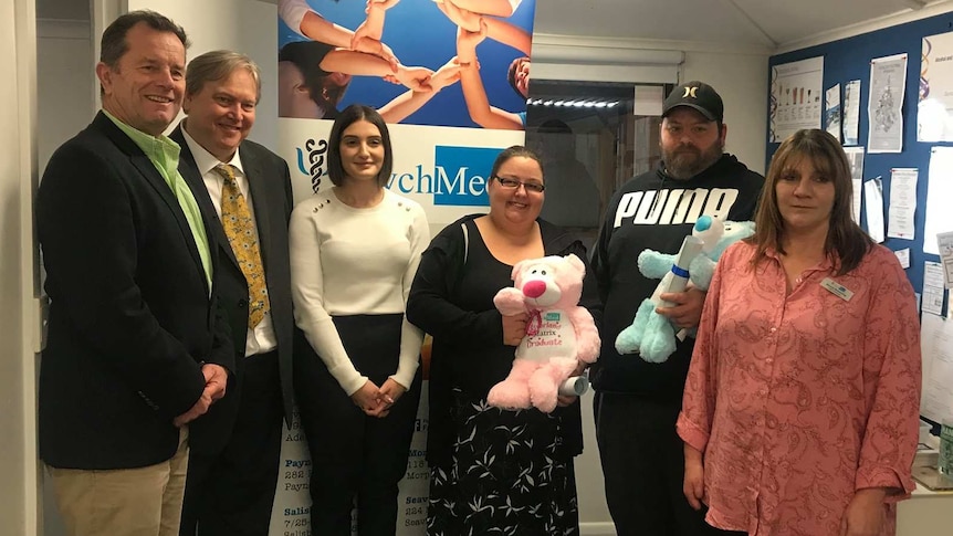 Six people stand in an office, two holding stuffed bears, at a drug treatment program graduation.