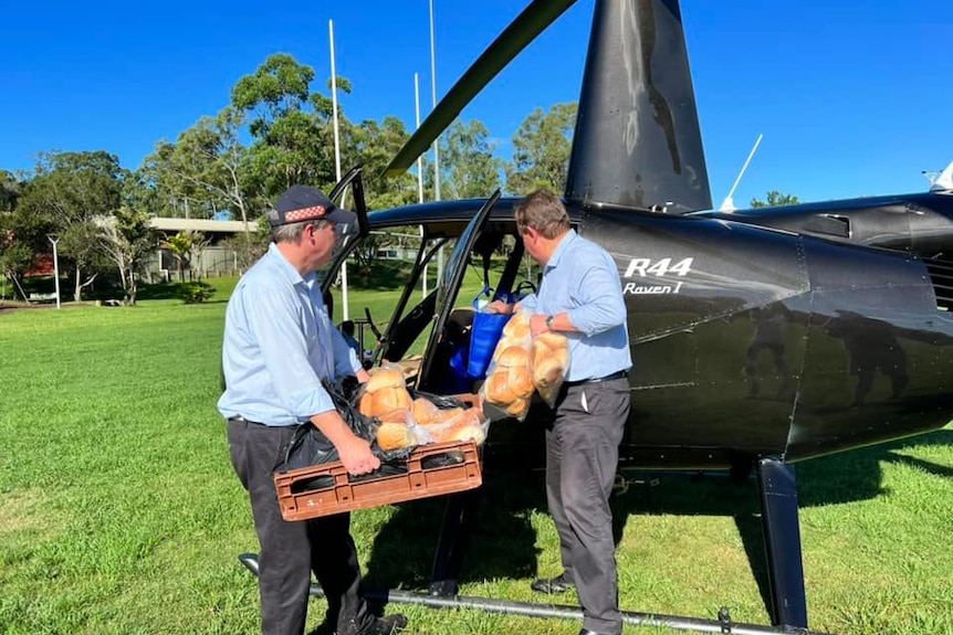 Two men load a pallet full of fresh bread into a black helicopter on a sports field. 