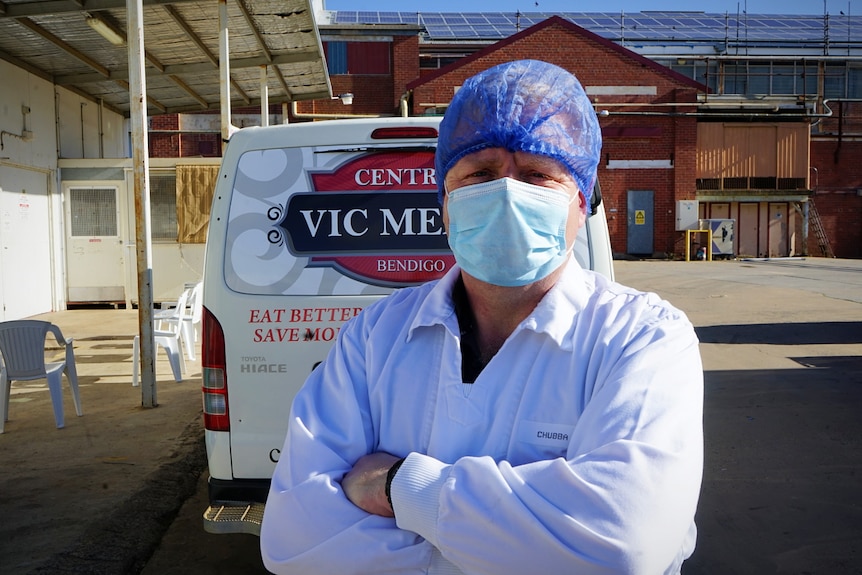 A man in a white smock and blue hair net stands in front of a van.