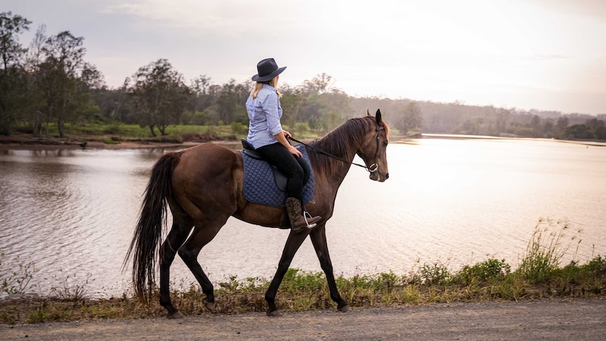 A woman rides a horse, with a river in the background, as the sun rises.