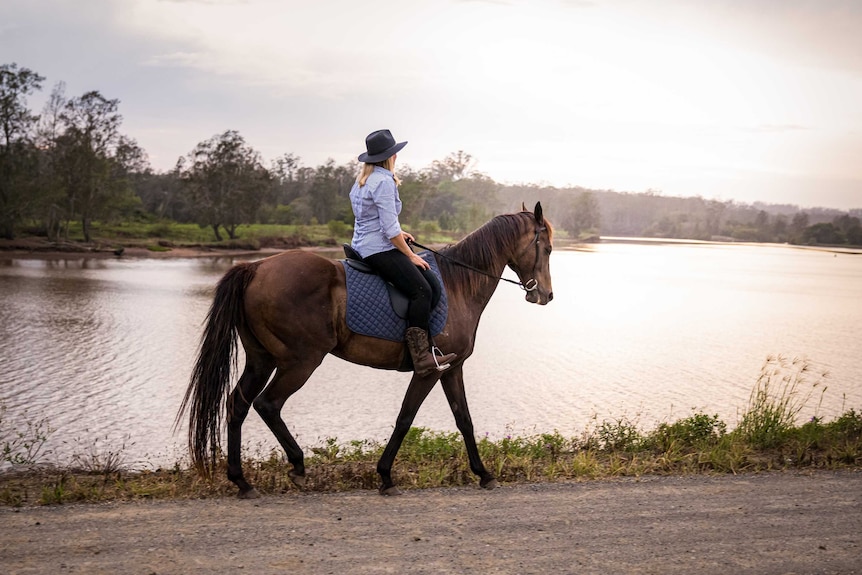 A woman rides a horse, with a river in the background, as the sun rises.