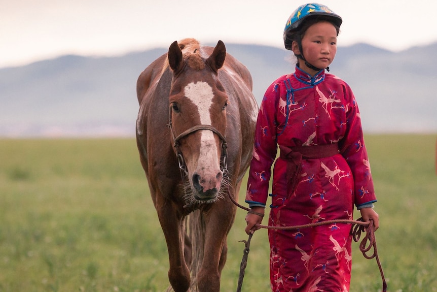 A young Mongolian girl walks her horse across the grass plains.