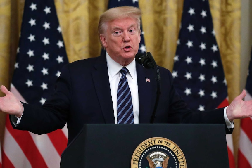 Donald Trump's arms are outstretched while speaking at a podium in front of American flags.