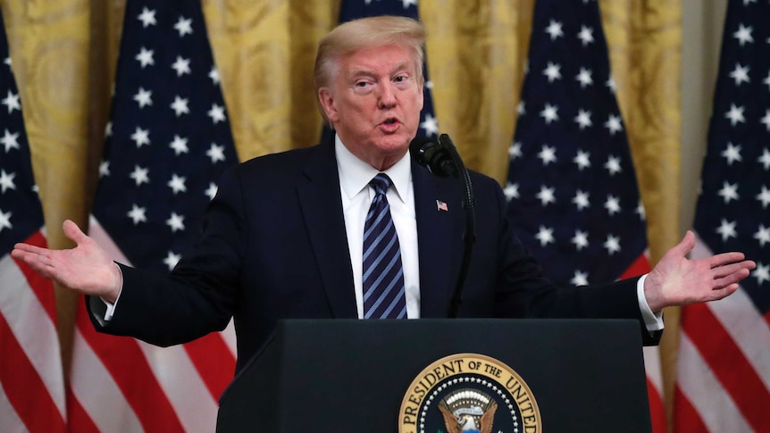 Donald Trump's arms are outstretched while speaking at a podium in front of American flags.