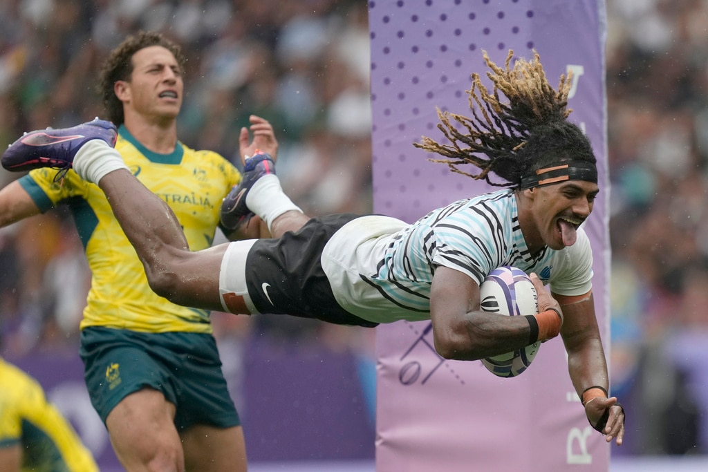 Fijian rugby player with his tongue poking out, dives through the air holding a rugby ball with tape round his head