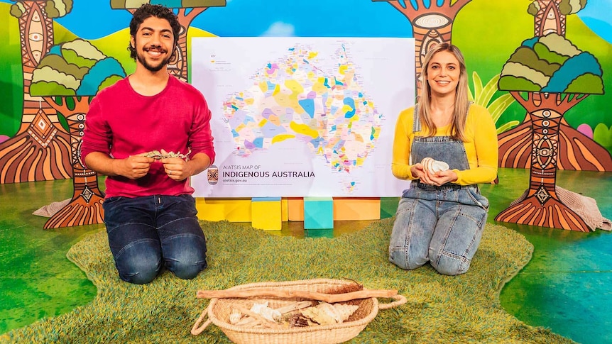 Hunter and Rachael holding leaves and a shell in front of a map showing the different regions of Indigenous Australia