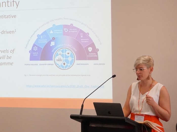 A woman with short blonde hair speaking at a lecturn with a powerpoint presentation behind her