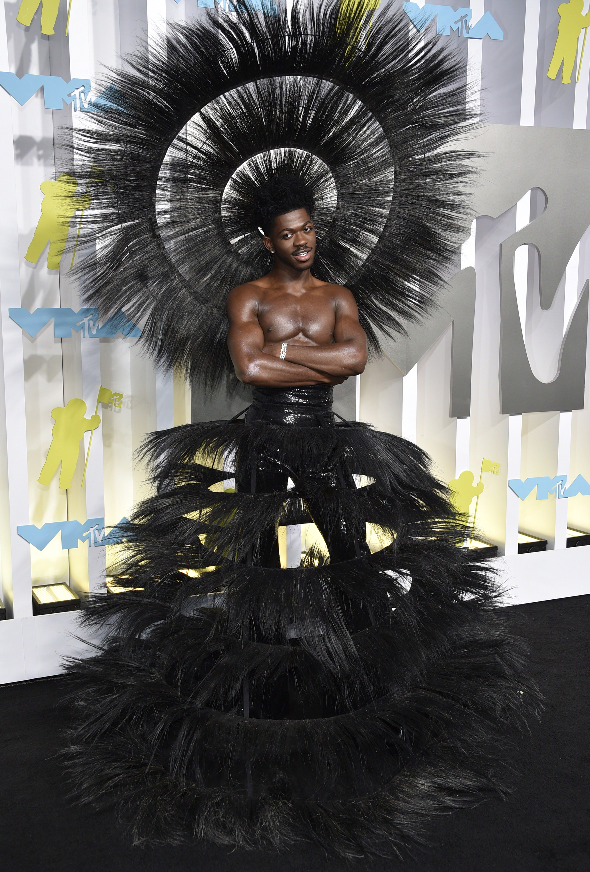 lil nas x poses with crossed arms on the vmas red carpet wearing a massive black spiky headpiece and black feathery skirt