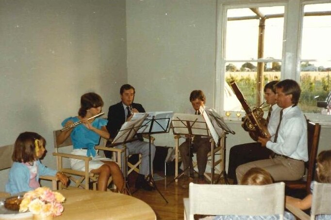 Five young adults in a white room play instruments in a semi-circle next to a window overlooking grassy fields.