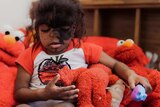 A young girl sits in a big pile of Elmo dolls in a play room.