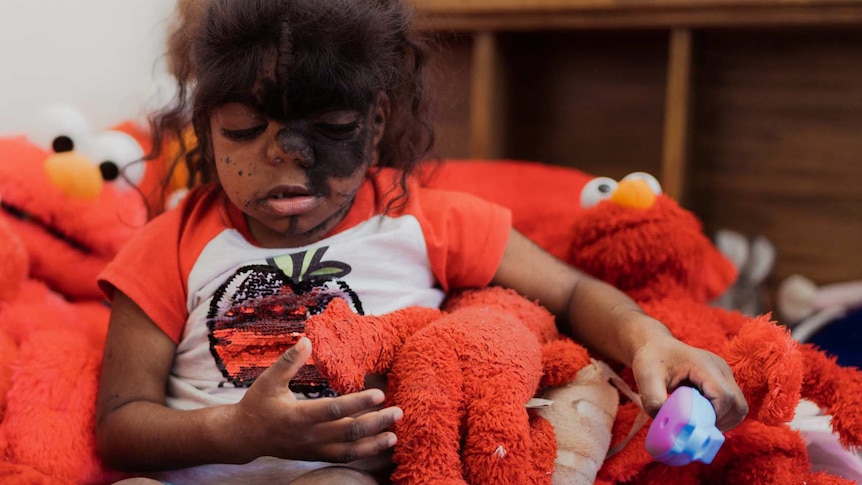 A young girl sits in a big pile of Elmo dolls in a play room.