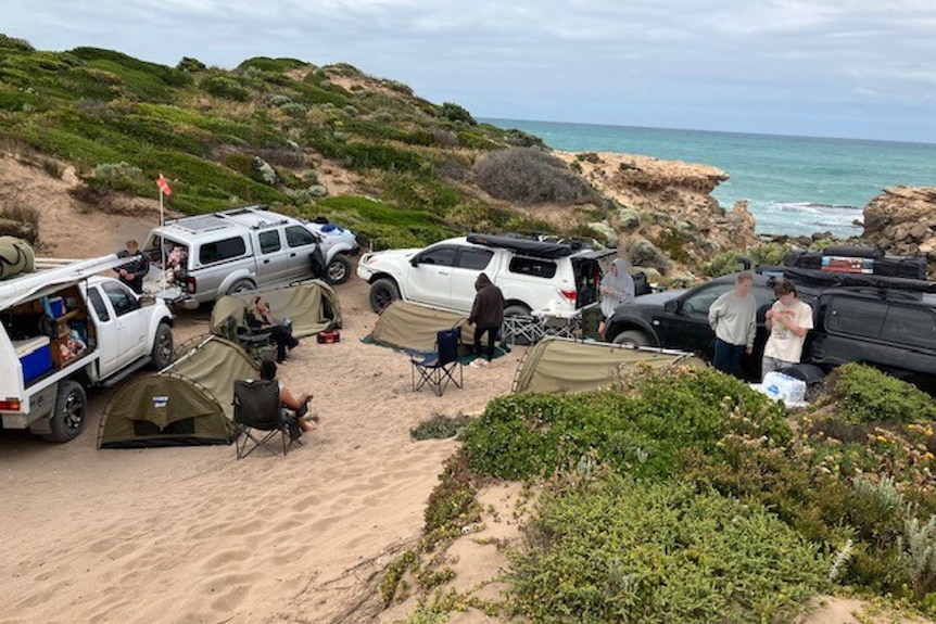 Four large SUV vehicles parked in a sand dune, a few swags pitched between them. A few people sit in camp chairs.