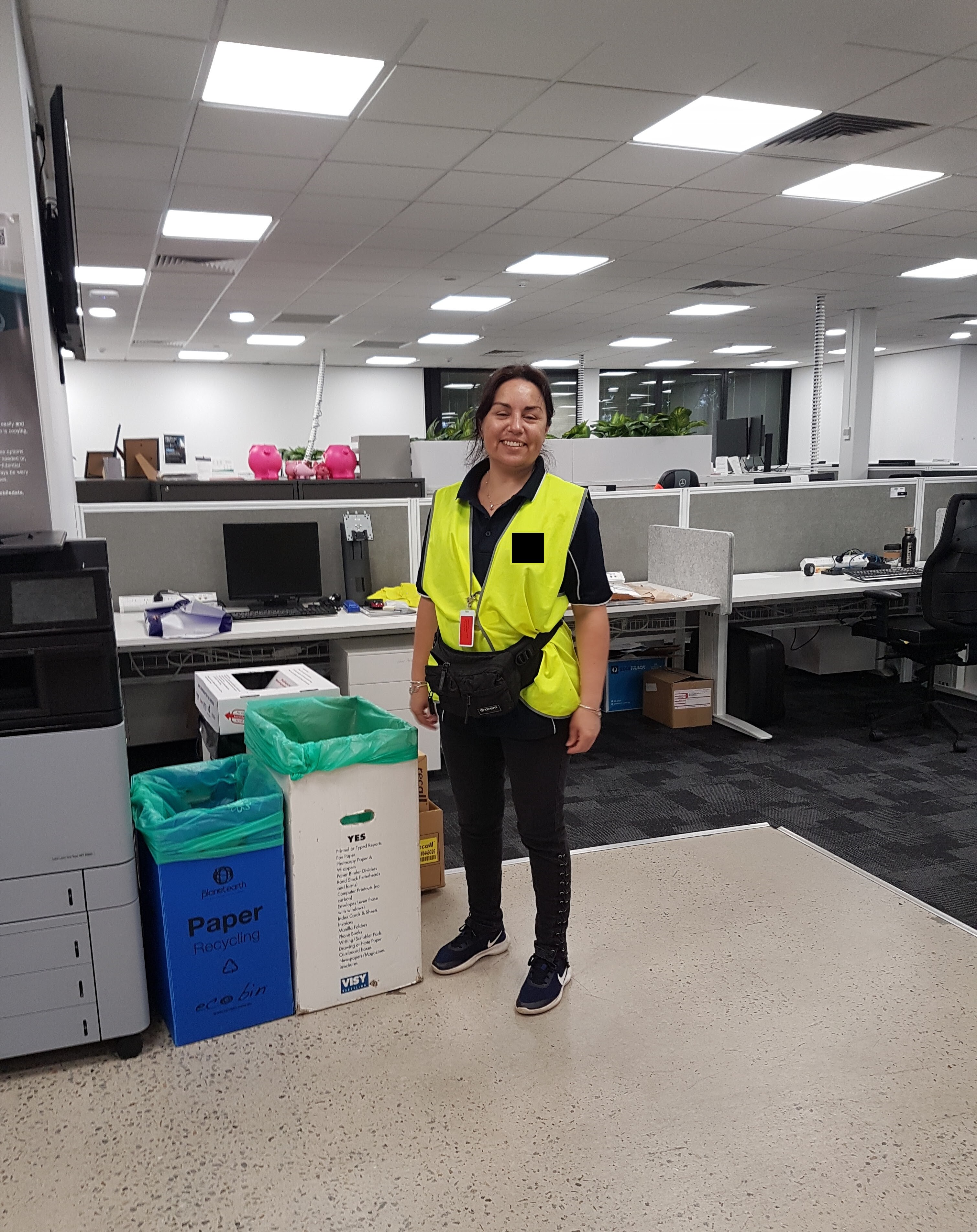 A woman wearing a bright yellow vest in an office stands next to paper bins and smiles.