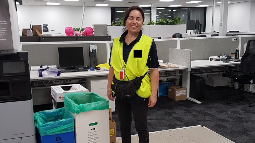 A woman wearing a bright yellow vest in an office stands next to paper bins and smiles.