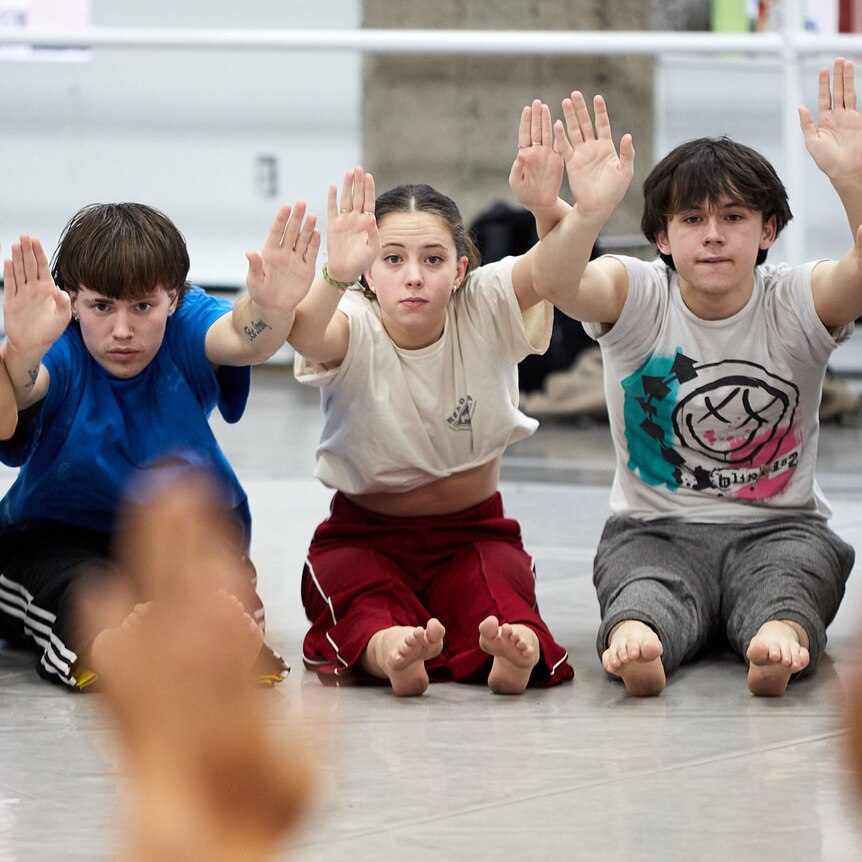 young people sit on a rehearsal room floor with legs and arms outstretched