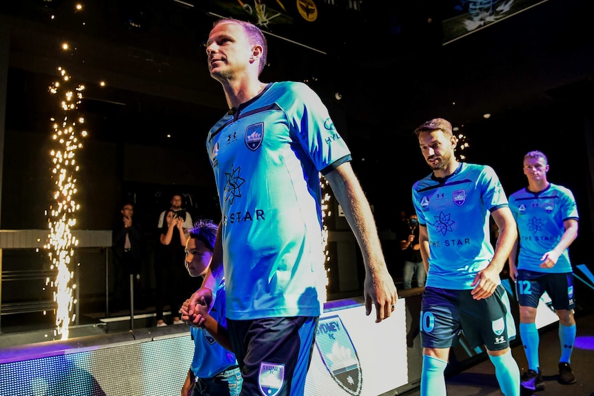 Three men in blue jerseys walk on a stage next to fireworks.