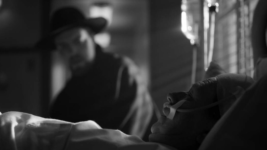 Black and white still showing in foreground Gary Oldman lying in hospital bed, and out of focus sitting beside him is Tom Burke.