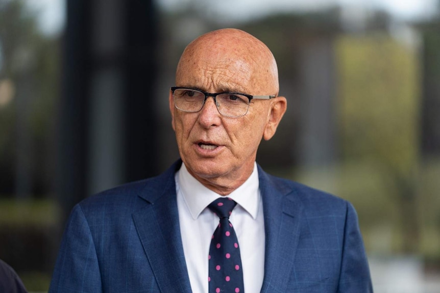 Un John Quigley à lunettes portant un costume bleu, une chemise blanche et une cravate bleue à pois roses.