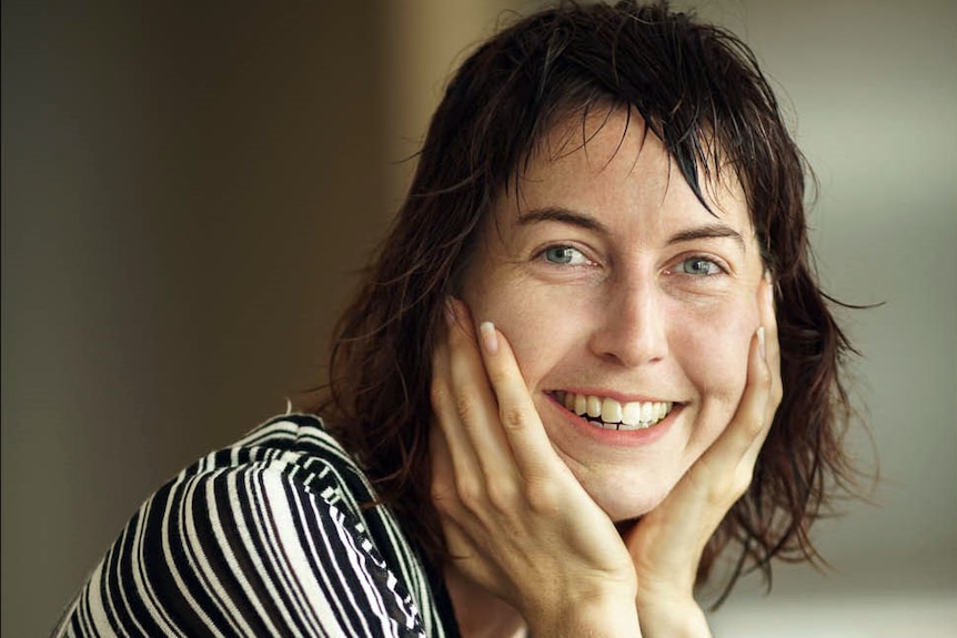 A woman smiles with her chin in her hands against a grey-green background.