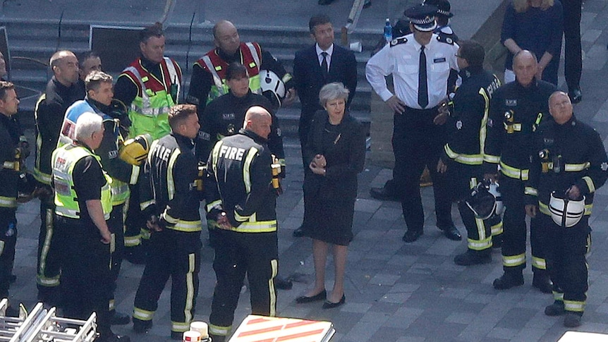 Theresa May stands near Grenfell Tower in London surrounded by emergency services members.