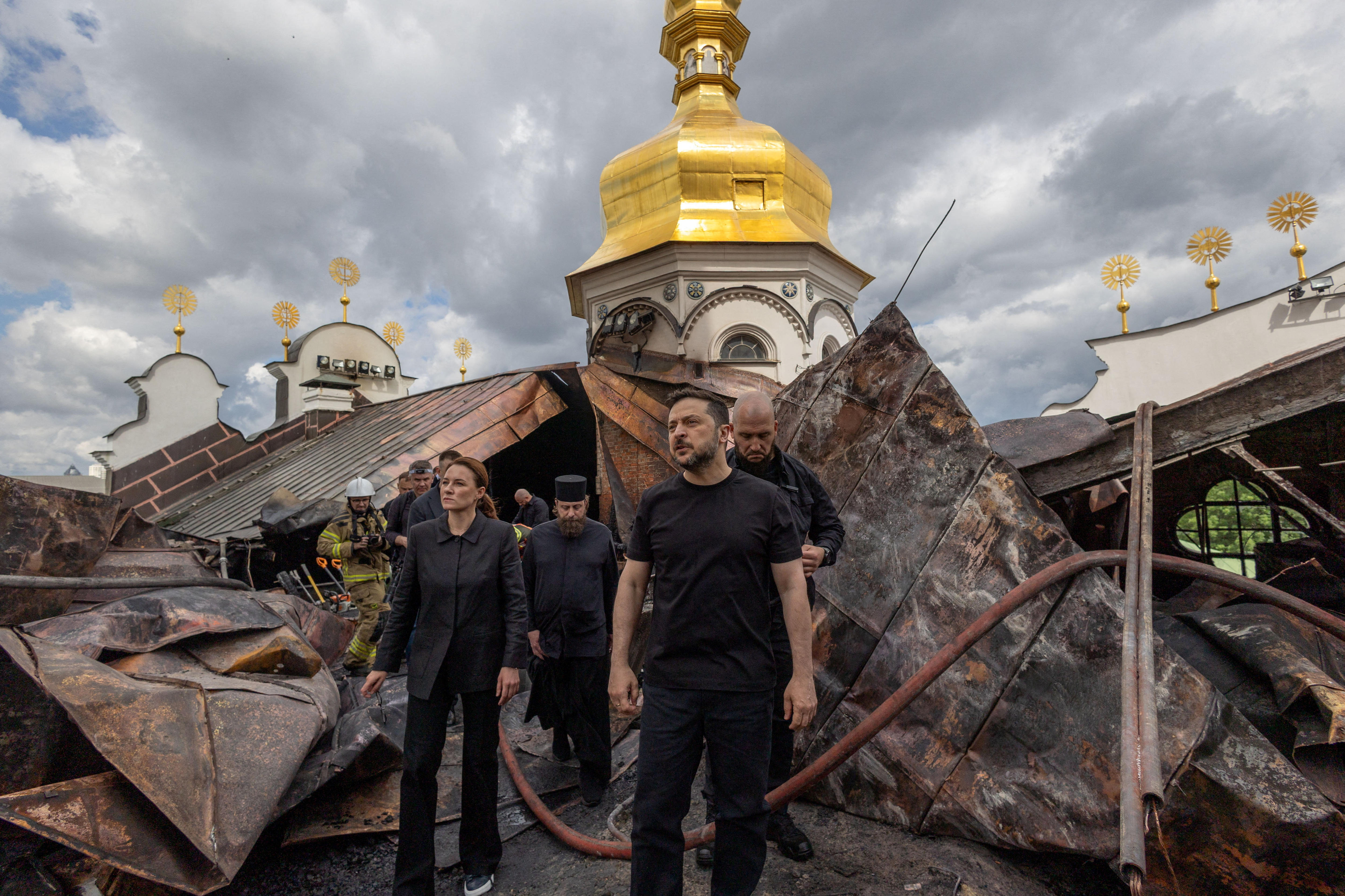 Three men and a woman dressed in black walk through charred ruins of a cathedral fire with golden dome of the complex behind.