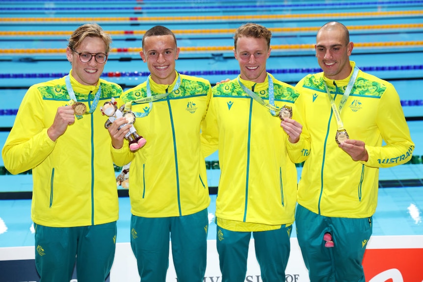 australian men's swim relay team smiles and poses with medals next to competition swimming pool