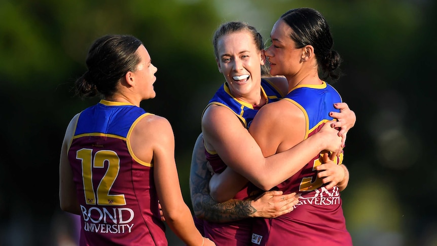 Two AFLW teammates embrace in celebration during a game, while a third teammate watches on.