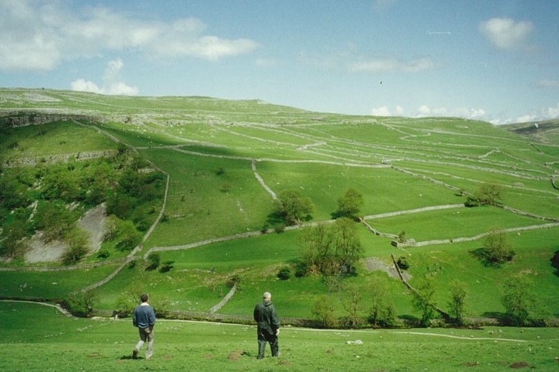 Two men walk towards green hill, lined with empty paddocks.