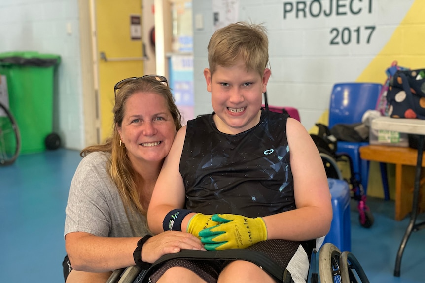 A mother crouches next to her son in a wheelchair and they both smile