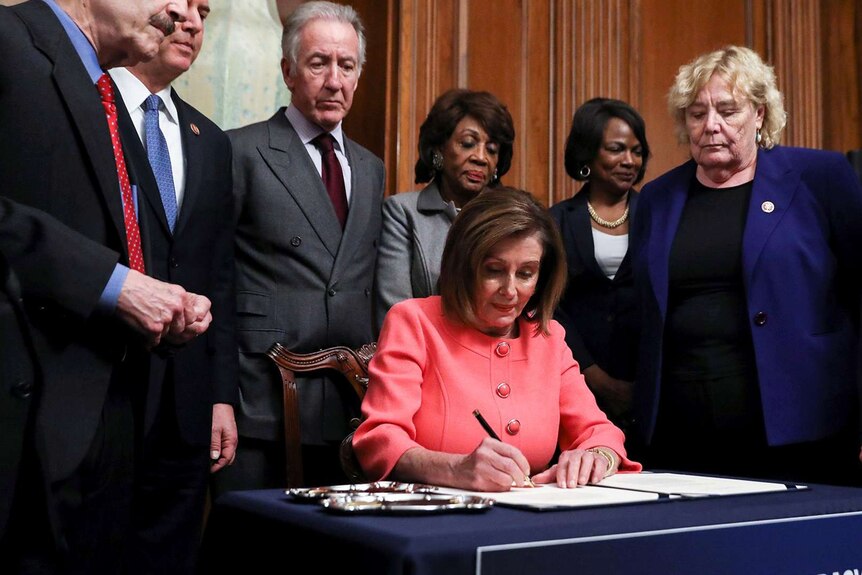 Nancy Pelosi sits at a desk, signing a document. A number of members of congress stand around her.