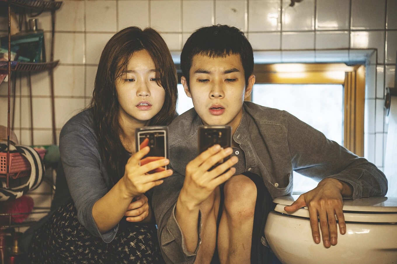 korean young male and female looking at mobile phones while crouched in a dirty tiled room, leaning against toilet bowl