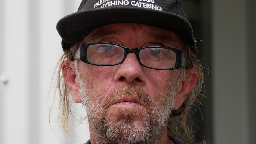 Up close shot of bearded man wearing black hat and glasses stares into camera