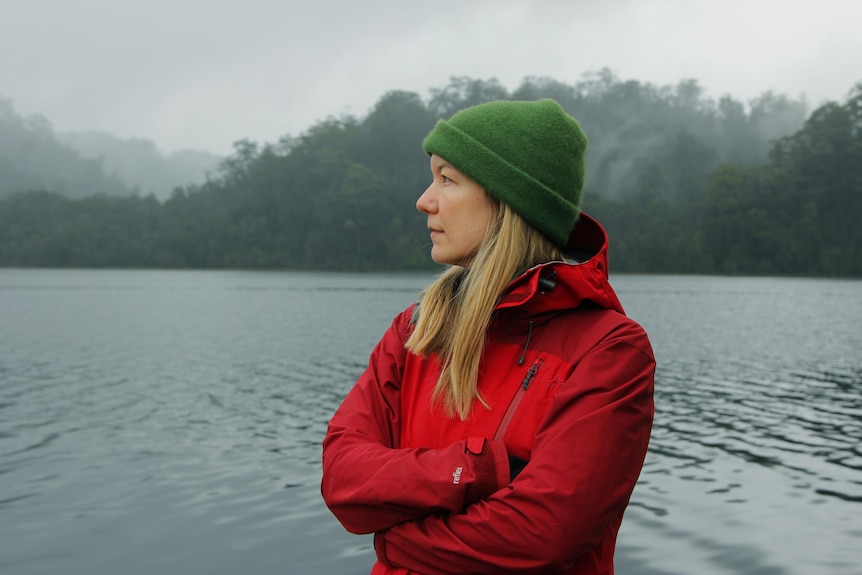 A woman wearing headphones and a red jacket stares off to the left of camera with a river in the background.