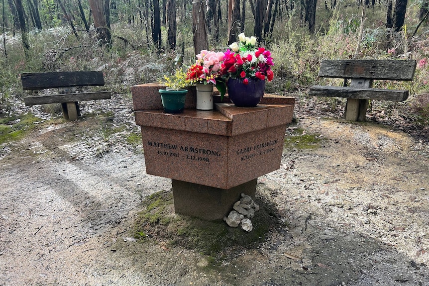 A marble square memorial with an inscription in the middle of a forest clearing with a lot of pots with colourful flowers. 