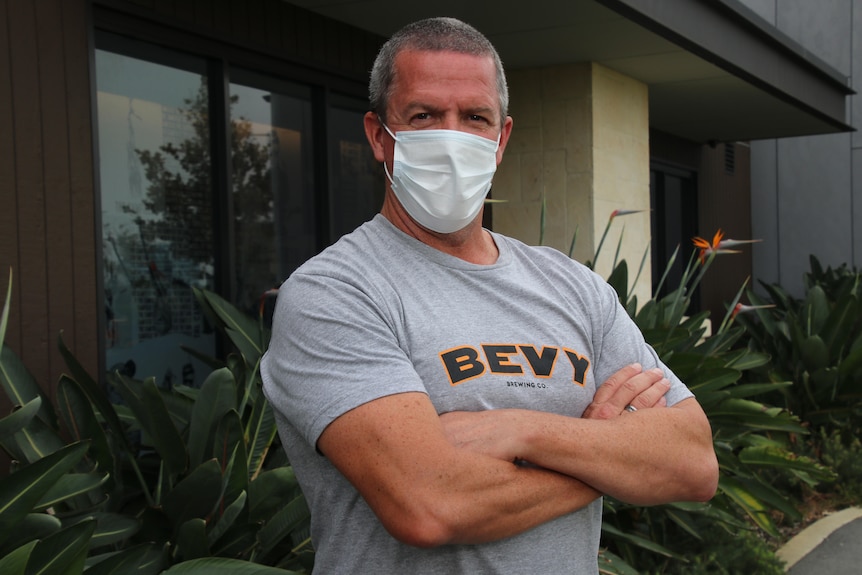 A mid-shot of a man wearing a face mask and a grey shirt posing for a photo with his arms folded.