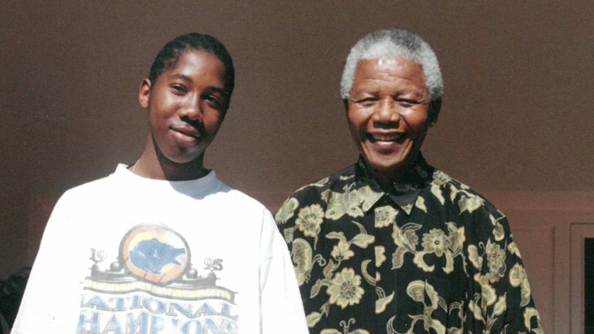 A teenager in white t-shirt next to man in black and yellow print shirt, with two boys standing in front of them. All smiling.