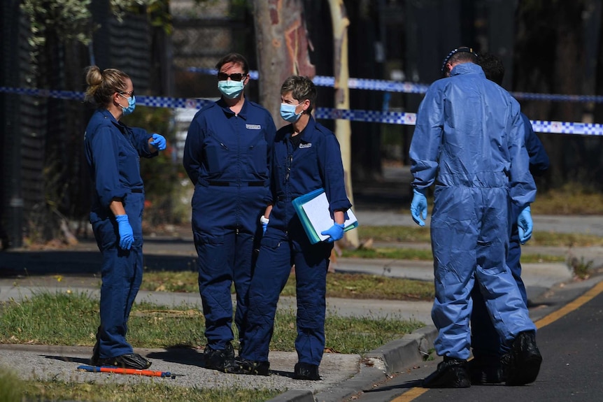 Four forensic police in blue jumpsuits and gloves at a crime scene.