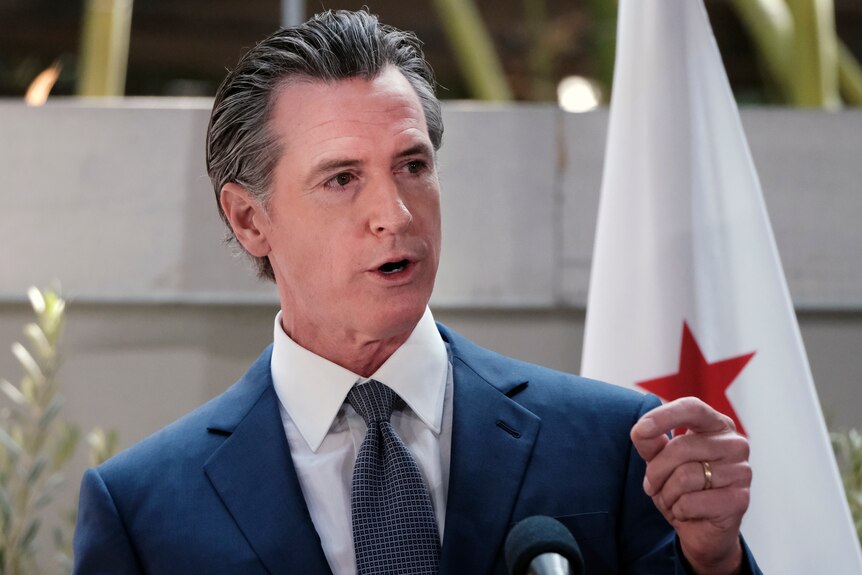 California Governor Gavin Newsom, wearing a jacket and tie and standing behind a podium, answers questions at a news conference