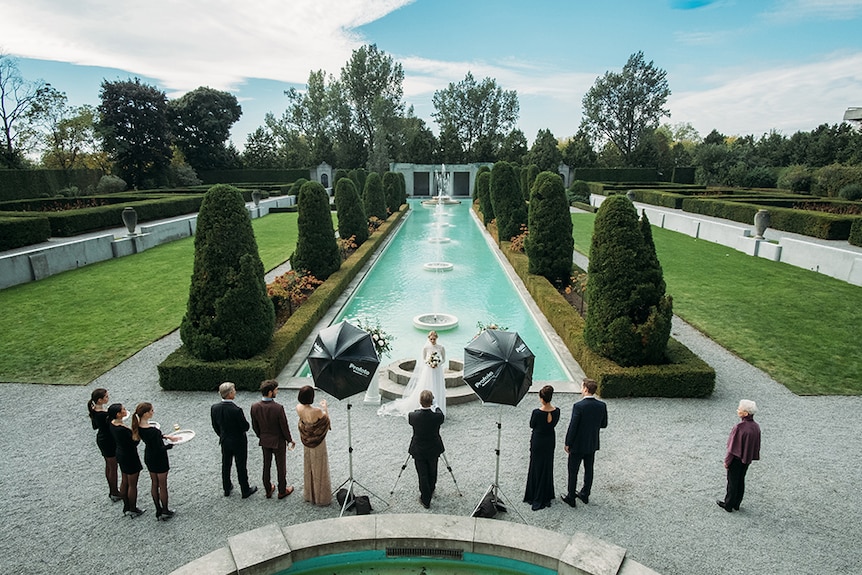 People watch as Samara Weaving poses for photo in front of long fountain in grand formal garden on a sunny day.