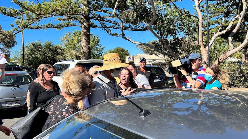 A woman screams at a man in a mask and hat as he gets into a car