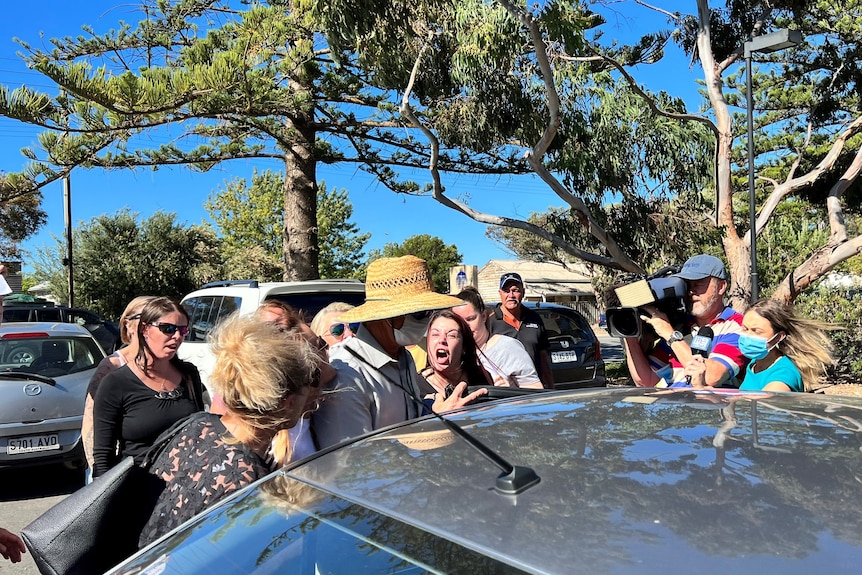A woman screams at a man in a mask and hat as he gets into a car