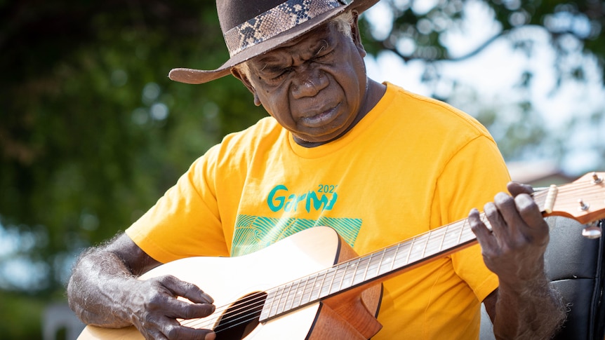 A man wearing a yellow shirt plays the guitar outside. He has a serious expression and looks at the frets.