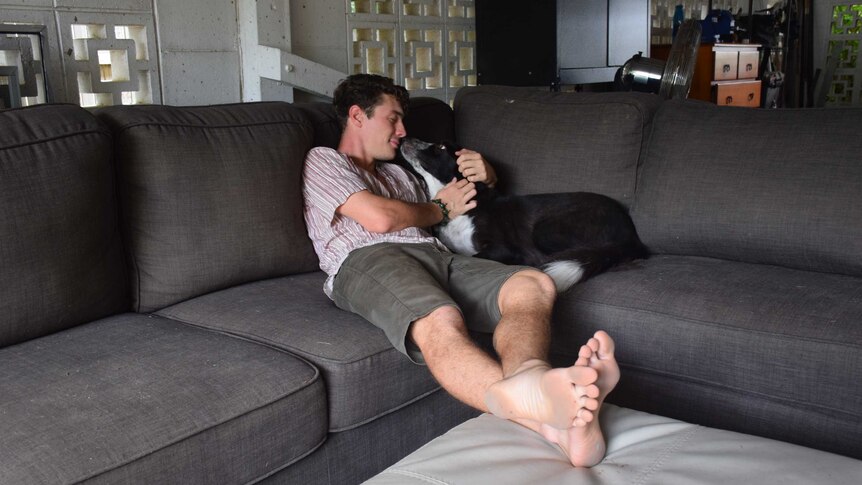 A man sits with his legs sprawled on a lounge. his Border Collie sits next to him and licks him on the mouth.
