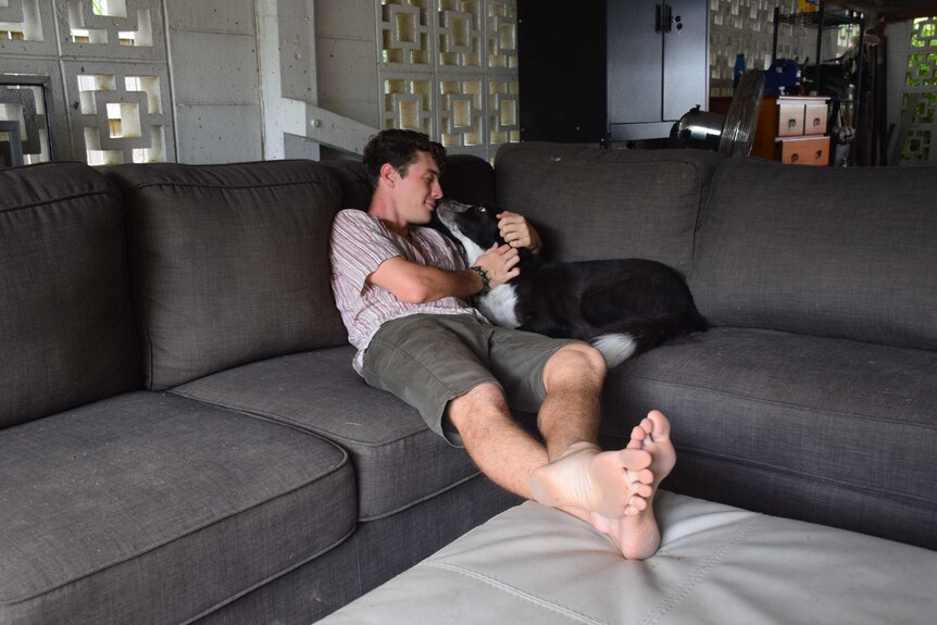 A man sits with his legs sprawled on a lounge. his Border Collie sits next to him and licks him on the mouth.