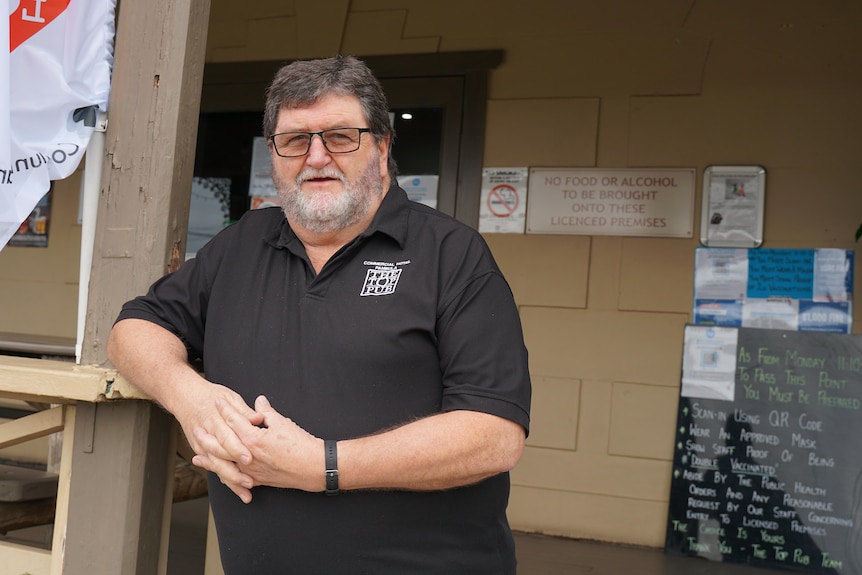 An older man with a beard and glasses stands outside his pub.