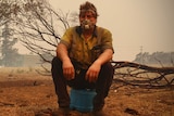 Luke Wright sits on an insulated water bottle wearing a face mask. His clothes are covered in ash and he's surrounded by smoke.