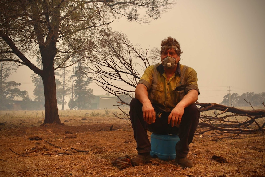 Luke Wright sits on an insulated water bottle wearing a face mask. His clothes are covered in ash and he's surrounded by smoke.